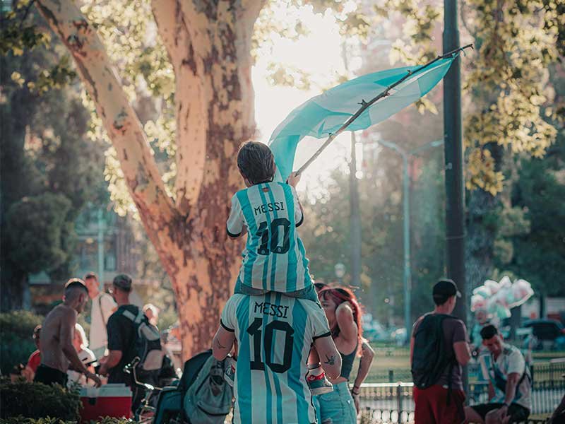 niño sobre los hombros de un hombre, levantando una bandera argentina, ambos con camiseta de futbol argentina que dice Messi