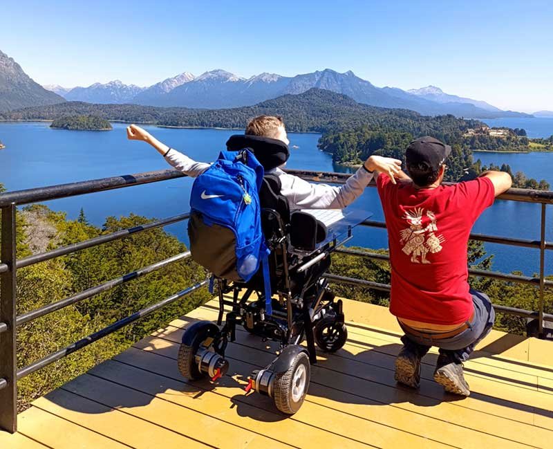 Persona usuaria de silla de ruedas y acompañante contemplando un paisaje de lago y montañas desde un mirador en Bariloche.